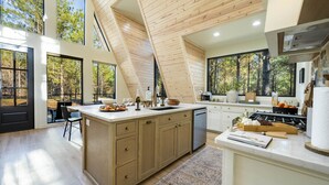A modern kitchen with a large island, wooden ceiling, and large windows offering a view of the forest.
