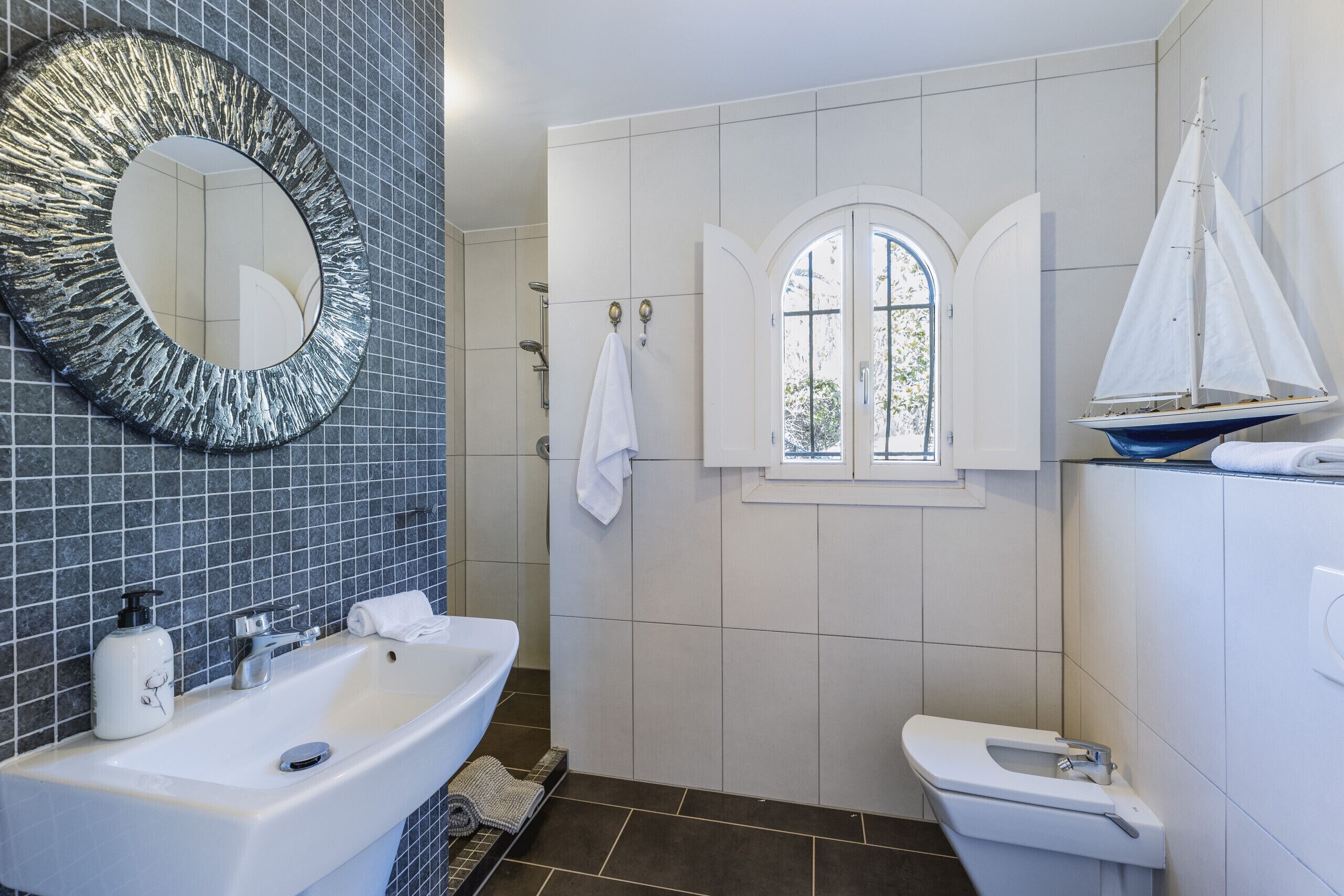 Un bany amb un mirall gran, un lavabo, un vàter i una petita finestra amb decoració de veler.