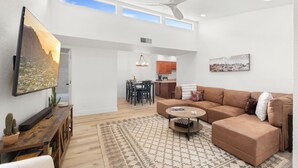 A spacious living room with a large flat-screen TV, a brown sectional sofa, a coffee table, and a patterned rug. There is a dining area with a table and chairs, and a kitchen area with wooden cabinets.