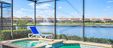 A pool area with a hot tub, a lounge chair, and a view of a lake and houses in the background.