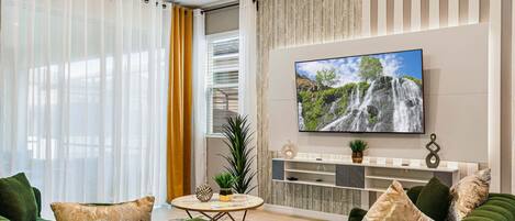 Sala de estar moderna com uma televisão grande, sofás verdes, uma mesa de centro e vista para uma cachoeira pela janela.