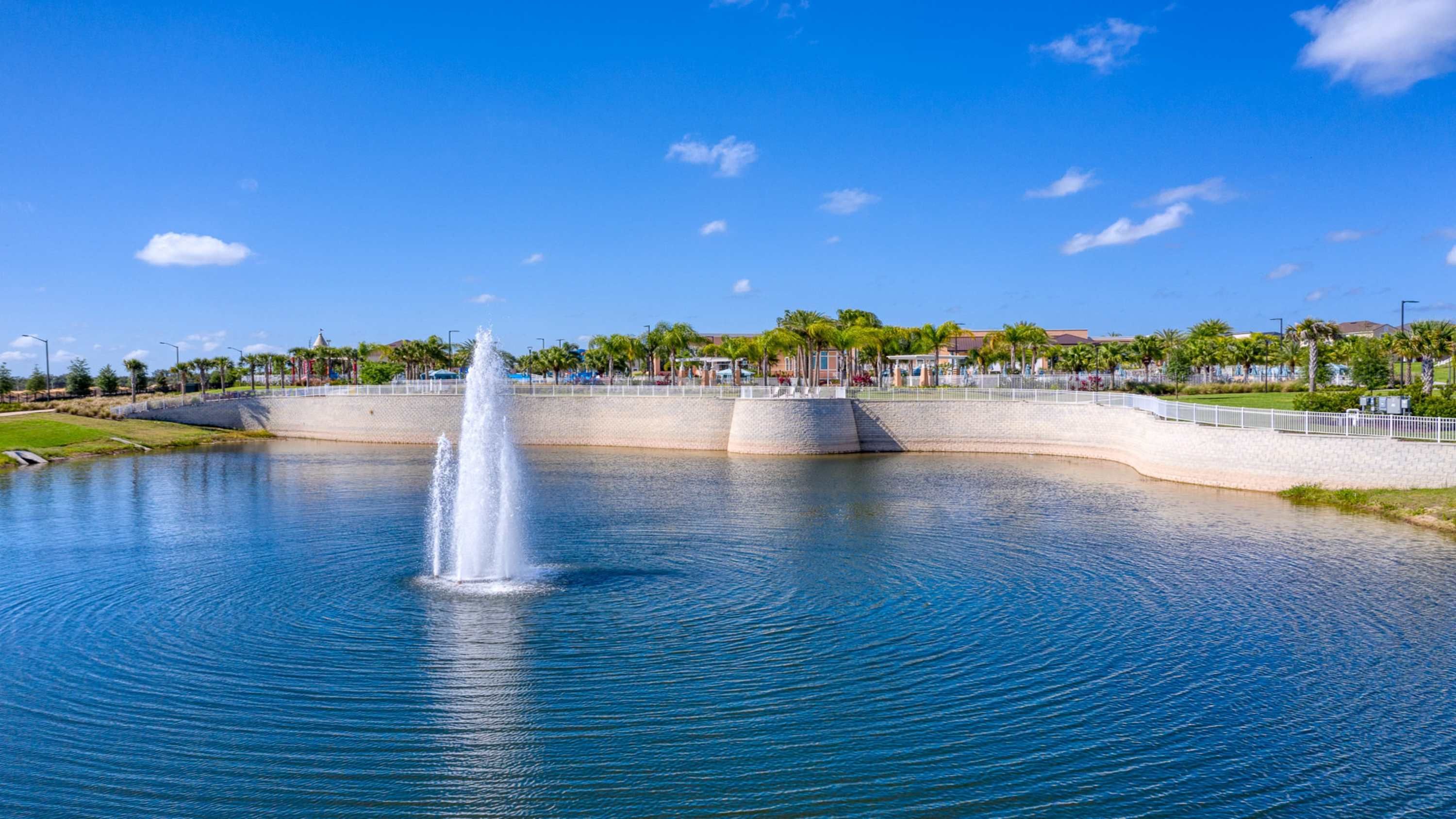 A large body of water with a central fountain, surrounded by a low stone wall and palm trees.