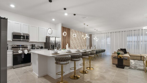 Una cocina moderna con armarios blancos, electrodomésticos de acero inoxidable y un salpicadero de mármol. Hay taburetes de bar con patas doradas y asientos tapizados en gris.