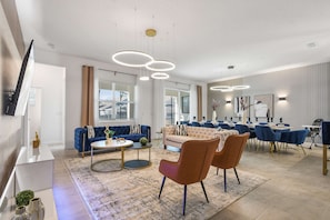 Un salon moderne comprenant un canapé, une table basse et des fauteuils, un coin repas avec des tables et des chaises, et un téléviseur à écran plat fixé au mur.
