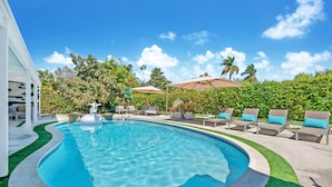 A swimming pool with a curved edge, surrounded by lounge chairs and umbrellas, with a view of palm trees and greenery.