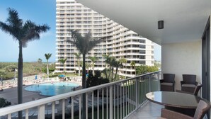Een balkon met een glazen tafel en rieten stoelen, met uitzicht op een zwembadgedeelte met palmbomen en een groot hotelgebouw op de achtergrond.