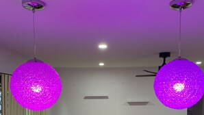 Una zona de menjador amb una taula de fusta i cadires blaves, dues làmpades penjants morades i un ventilador de sostre.