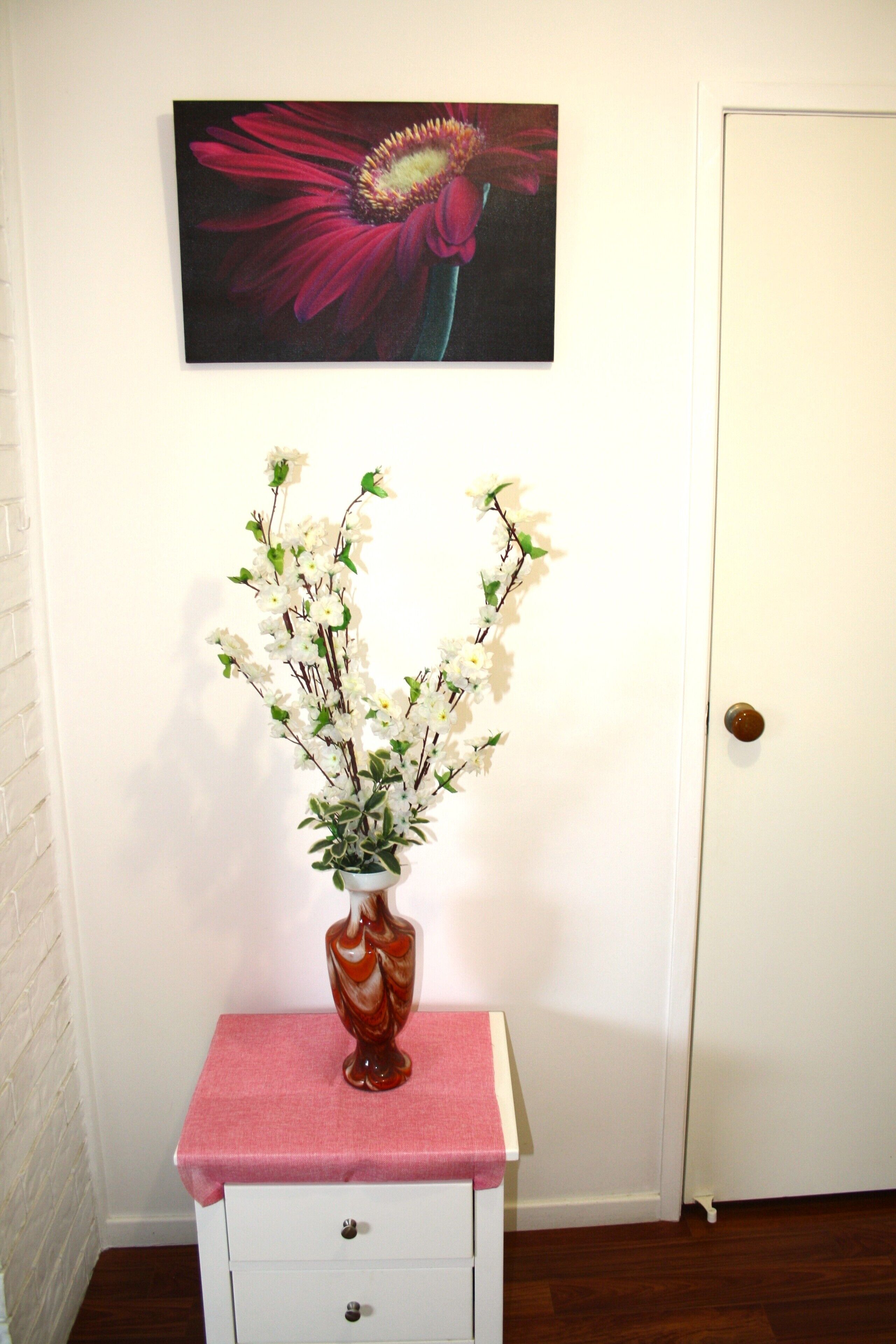 A white dresser with a vase of white flowers on top, a pink cloth, and a painting of a flower on the wall.