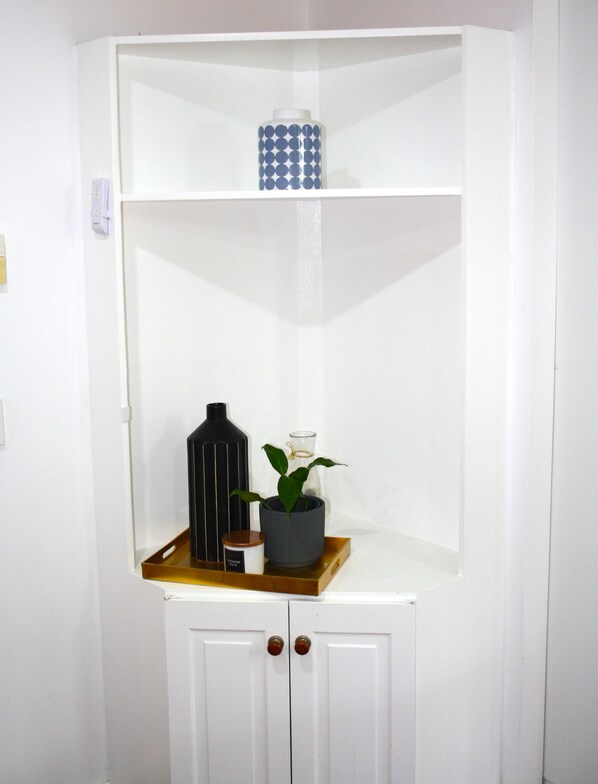A white corner cabinet with two shelves, a black vase, a potted plant, and a small container.