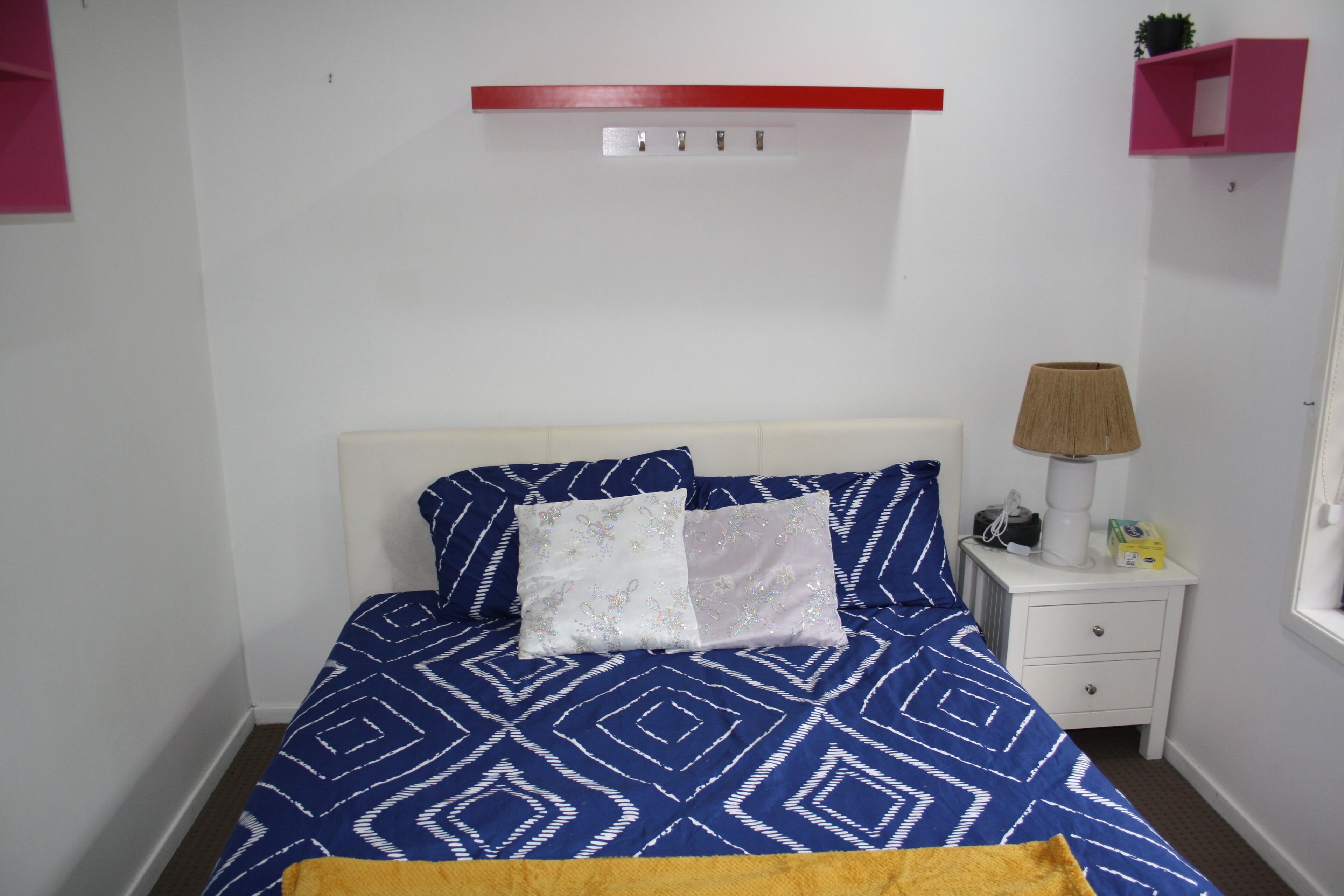 A bed with a geometric patterned bedspread, a white headboard, and two decorative pillows. There is a bedside table with a lamp and a small plant.