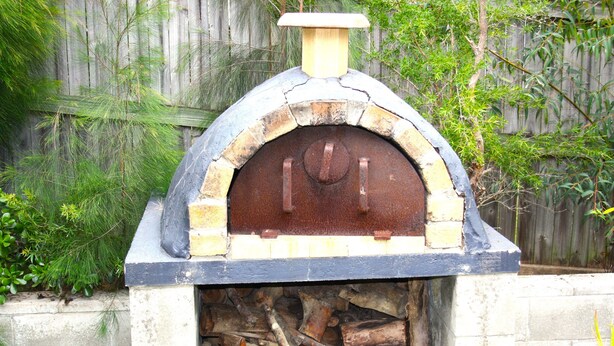 Forn de pizza exterior de maó amb una xemeneia, envoltat d'una tanca de fusta i vegetació.