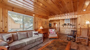 Interior acogedor de una cabaña con techo de madera, un sofá con un cojín que dice 'HOGAR', una mesita de centro y un sillón mecedora.