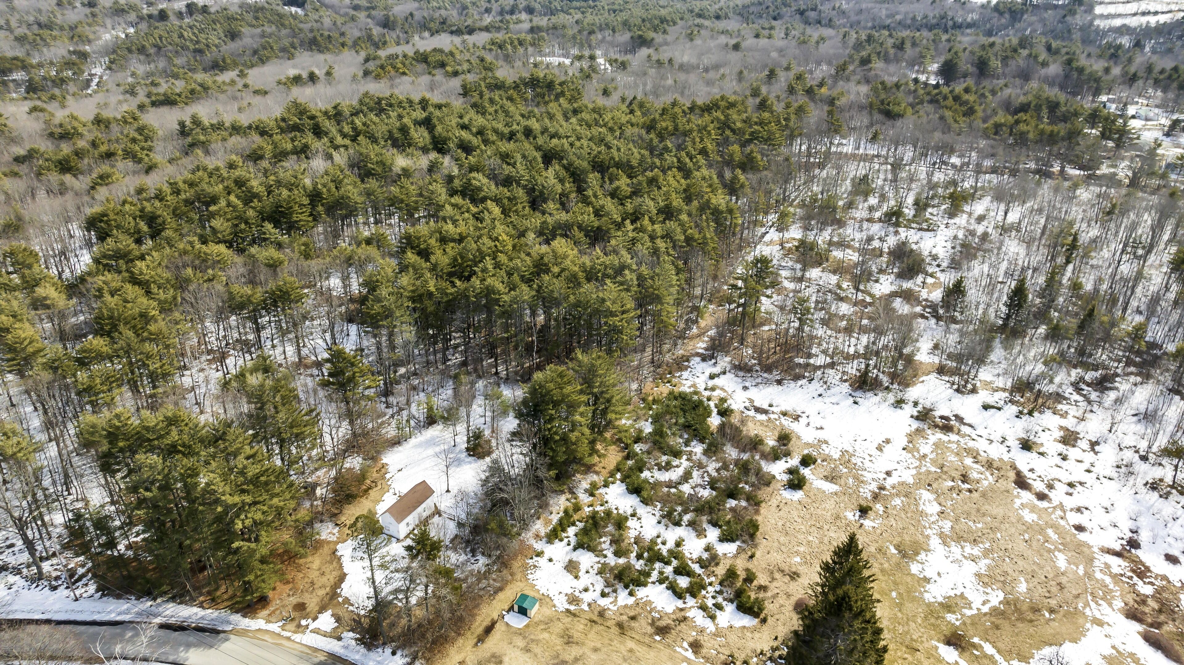 An aerial view of a forested area with scattered snow patches and a small building.
