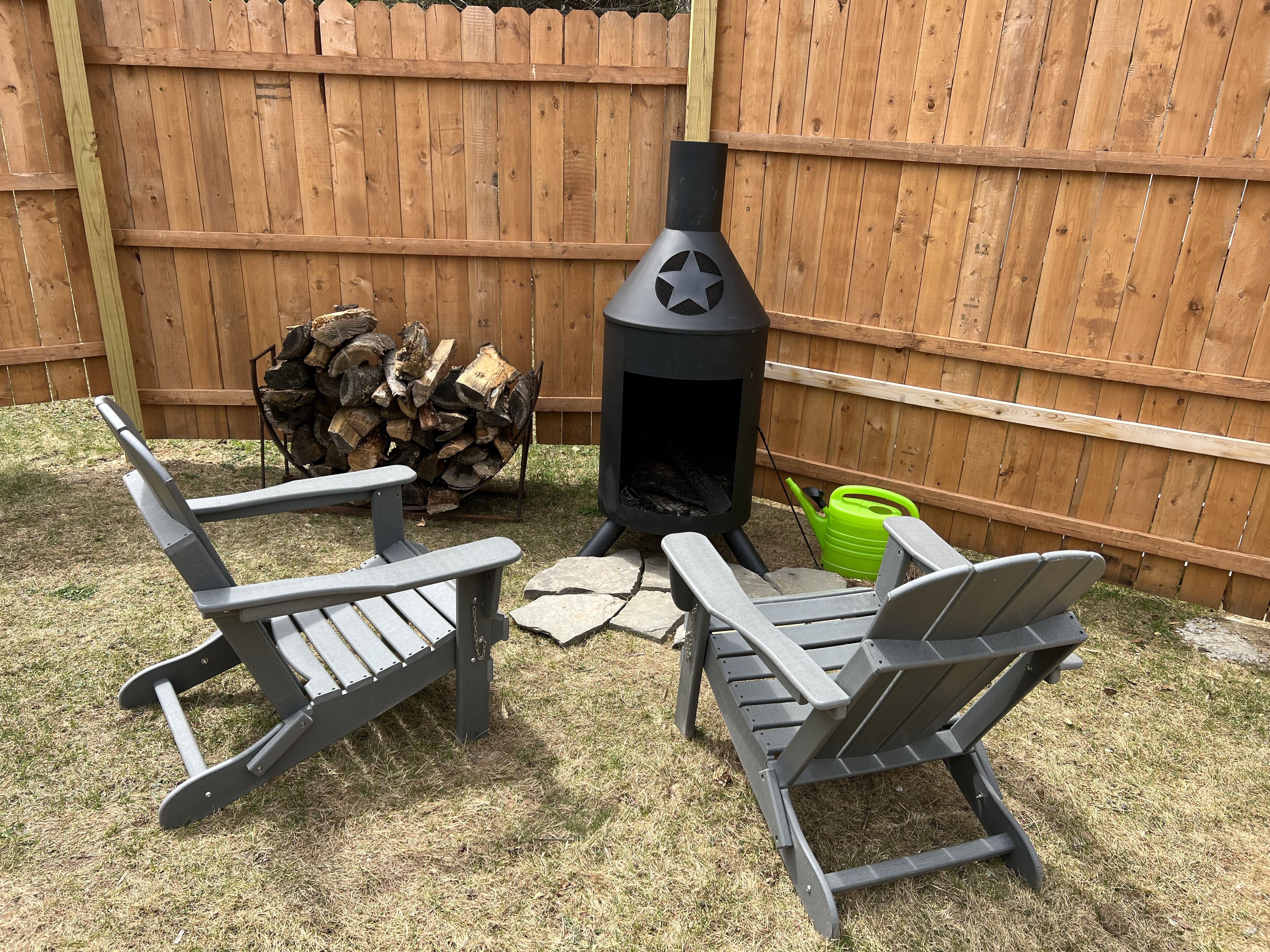A backyard with a wood-burning stove, two Adirondack chairs, a stack of firewood, and a green watering can.