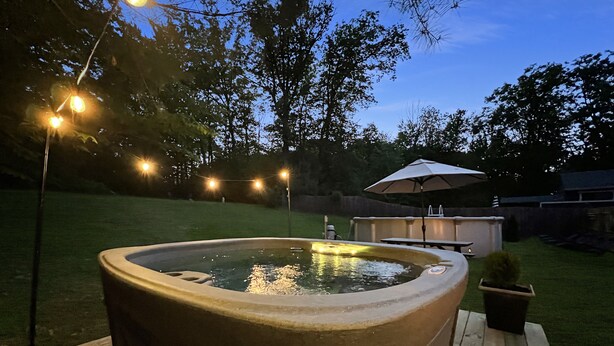 Ein Whirlpool auf einer Holzterrasse mit Sonnenschirm und Lichterkette in einem Garten.