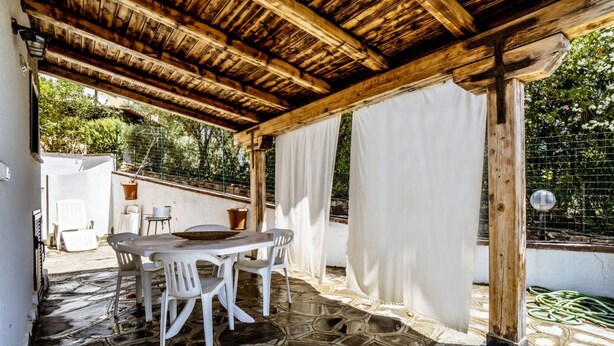 An outdoor patio with a stone floor, a wooden pergola, white curtains, and a table with chairs. - Casa Vacanza La Collinetta | 2 bedroom, 2 baths, veranda (budoni)
