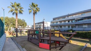 Un parque infantil con estructuras de madera y un tobogán amarillo, rodeado de palmeras y un edificio con balcones.