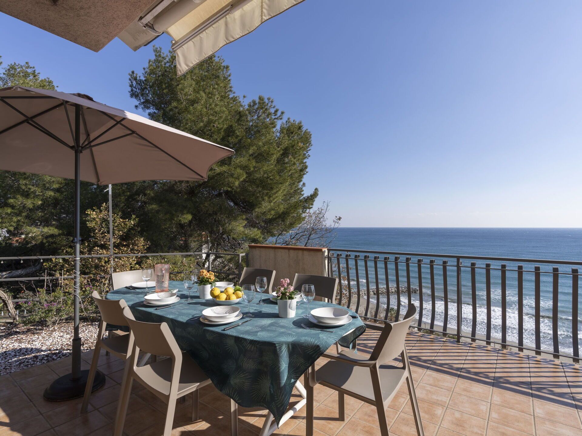 Mesa para dos con vista al mar, un patio con barandilla y un árbol al fondo.
