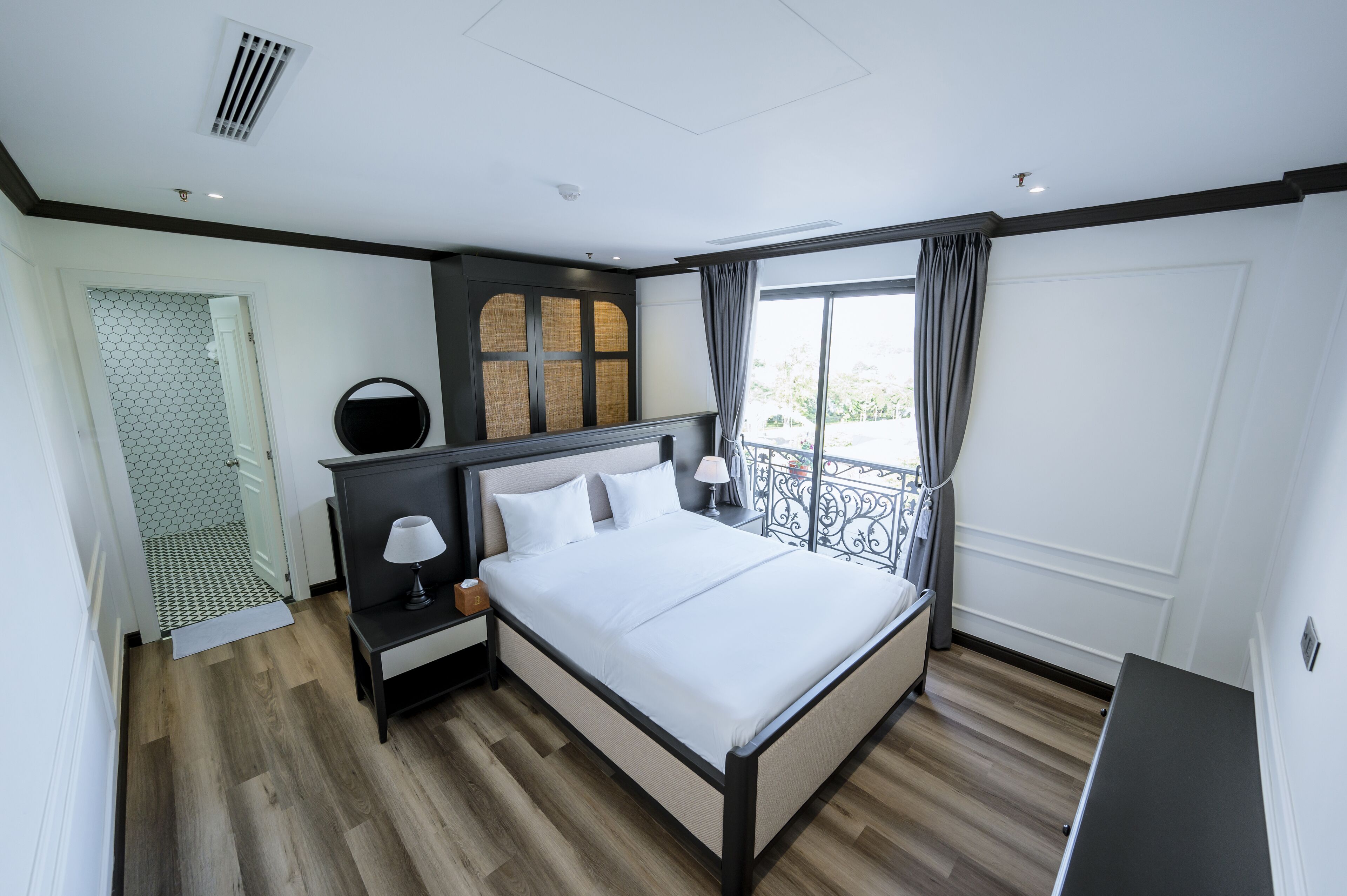 A modern hotel room with a large bed, a nightstand with a lamp, a mirror, and a view of the outdoors through a balcony.