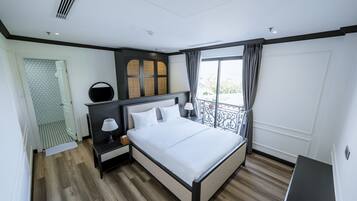A modern hotel room with a large bed, a nightstand with a lamp, a mirror, and a view of the outdoors through a balcony.