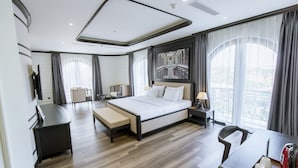 A modern hotel room with a large bed, a desk, a chair, and a view of the city through large windows.