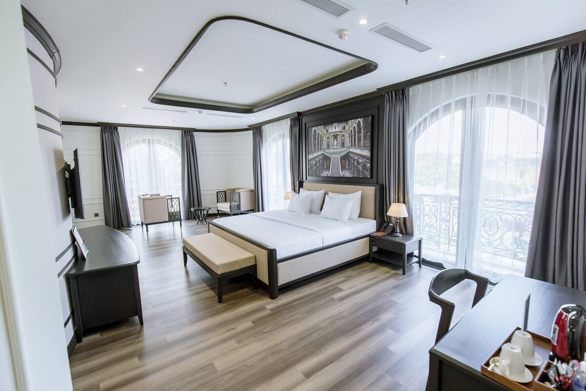 A modern hotel room with a large bed, a desk, a chair, and a view of the city through large windows.