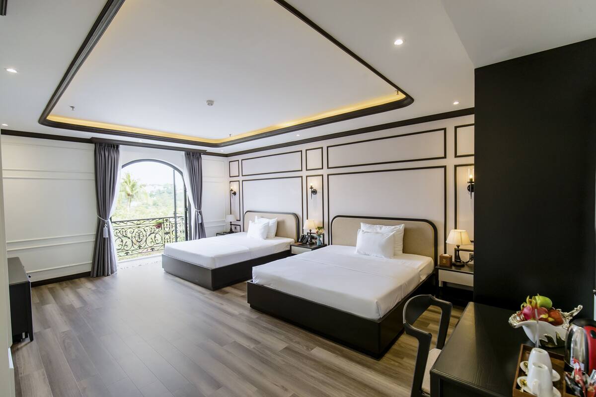 A hotel room with two beds, a balcony with curtains, a wooden floor, a desk with a tray of items, and a view of greenery outside.