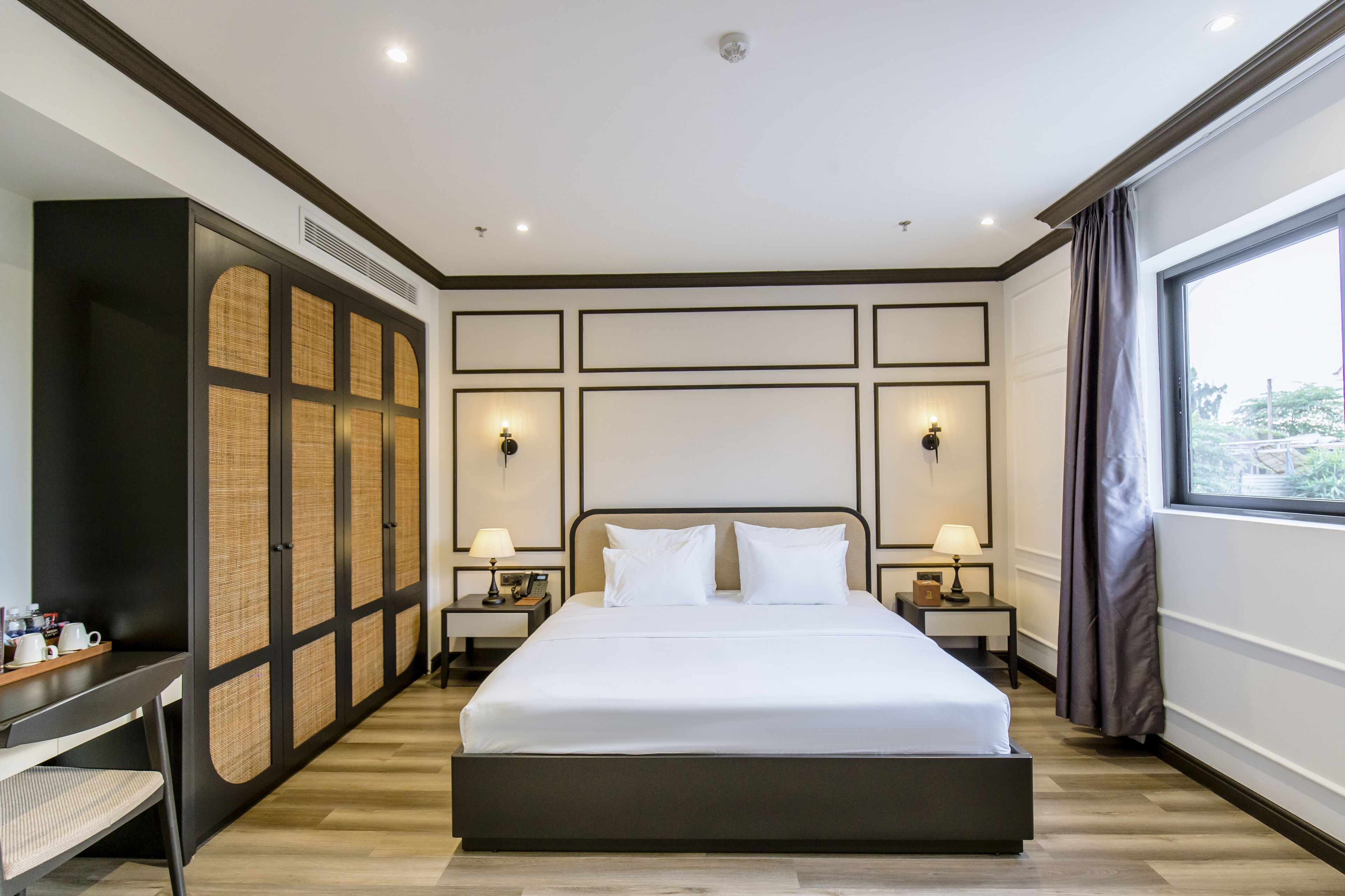 A modern hotel room with a large bed, two bedside tables with lamps, a wardrobe with a woven panel design, and a large window with curtains.