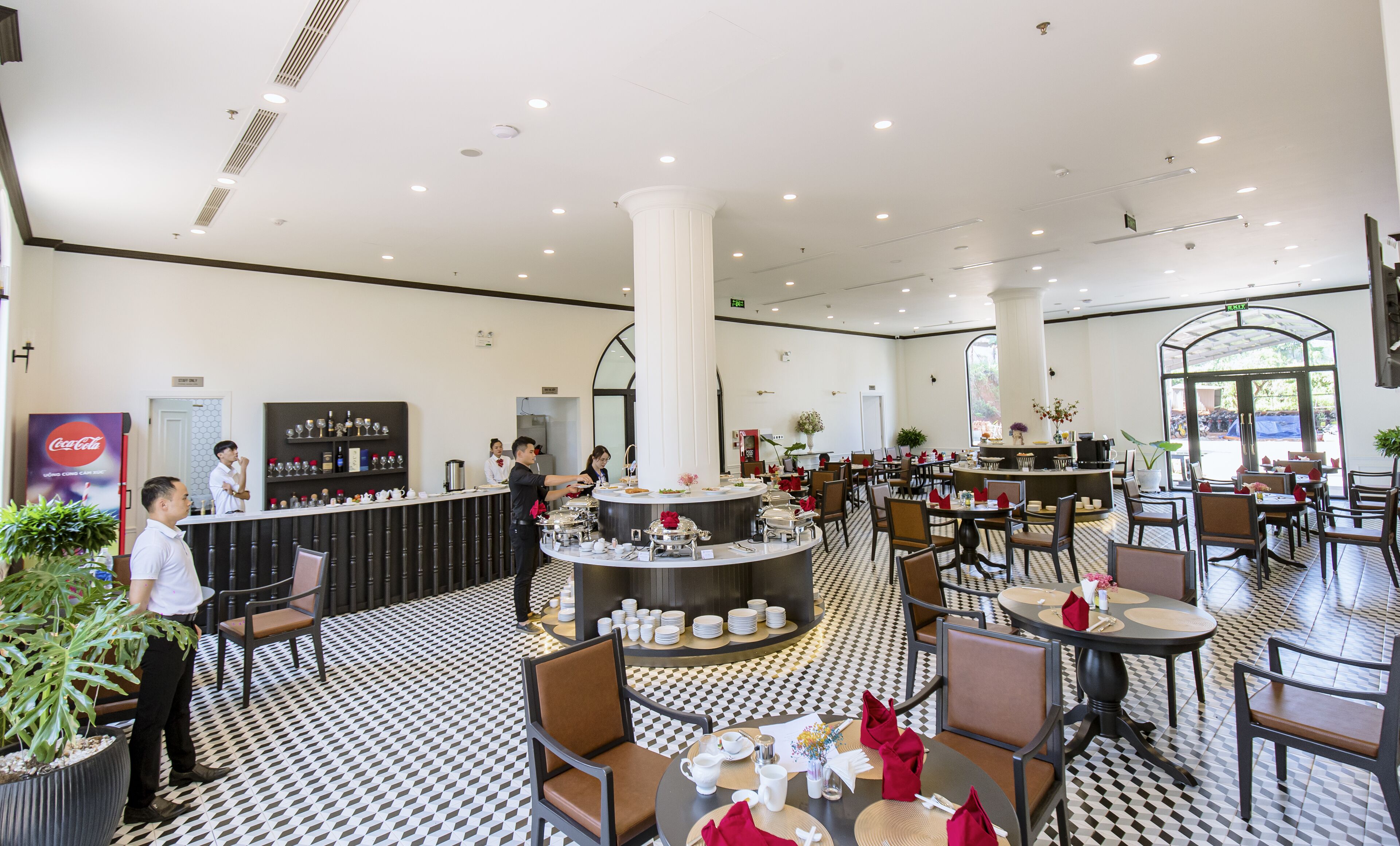 A spacious hotel lobby with a central bar, multiple seating areas, and a patterned floor.