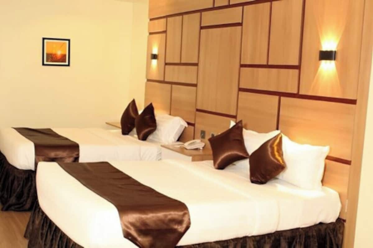 Een hotelkamer met twee bedden, elk met een bruin hoofdbord, witte beddengoed en een bruine plaid. Er zijn wandlampen en een lijst met een afbeelding aan de muur.