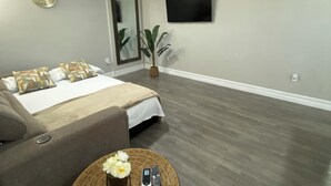 Une chambre d’hôtel moderne comprenant un lit, un canapé, une table ronde en osier avec un téléphone et des télécommandes, un téléviseur fixé au mur et une horloge.