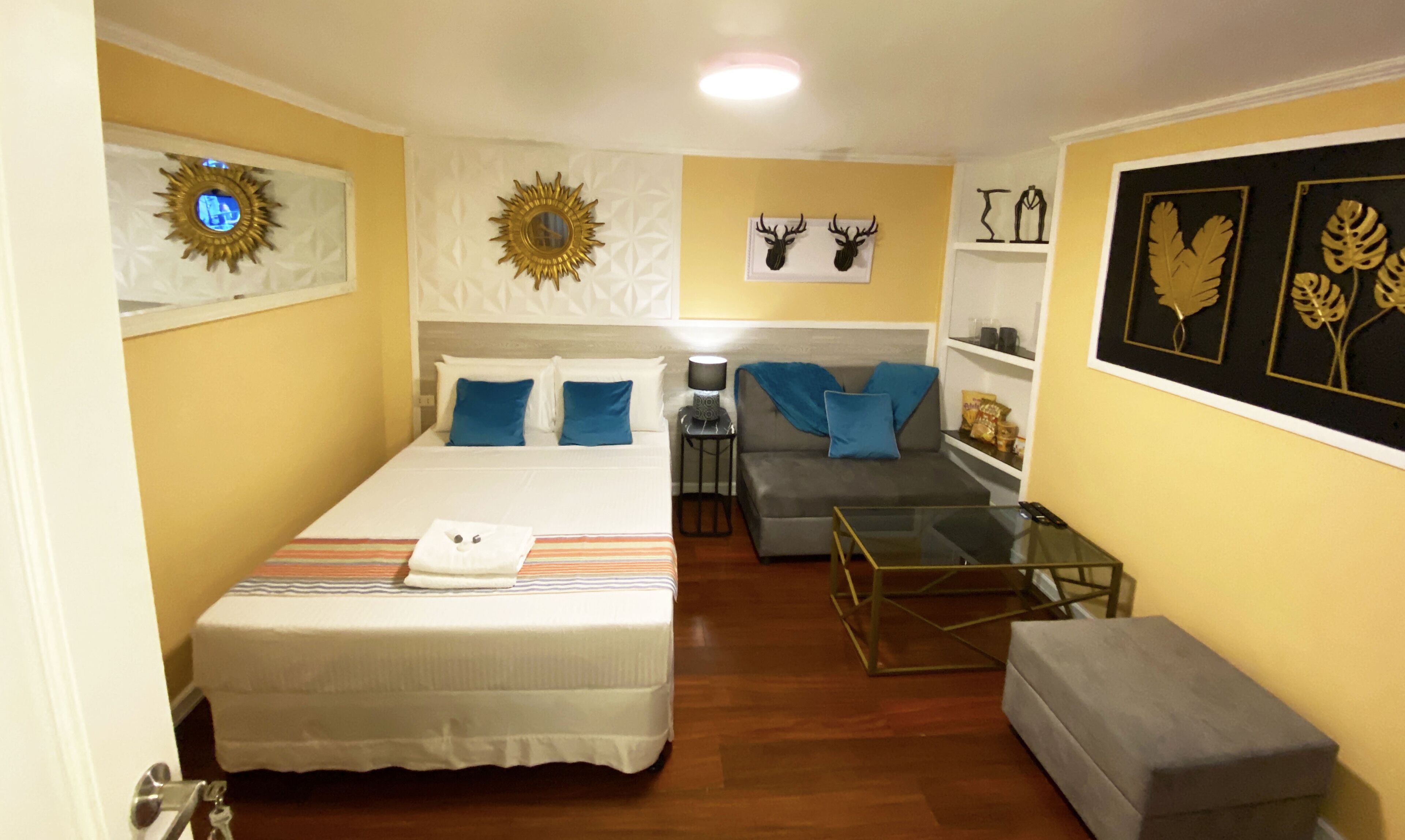 Habitación de hotel con cama, sofá, una mesita de centro de cristal y una mesita con lámpara. Decoración de pared que incluye un espejo con diseño de sol, monturas de ciervo y hojas enmarcadas.