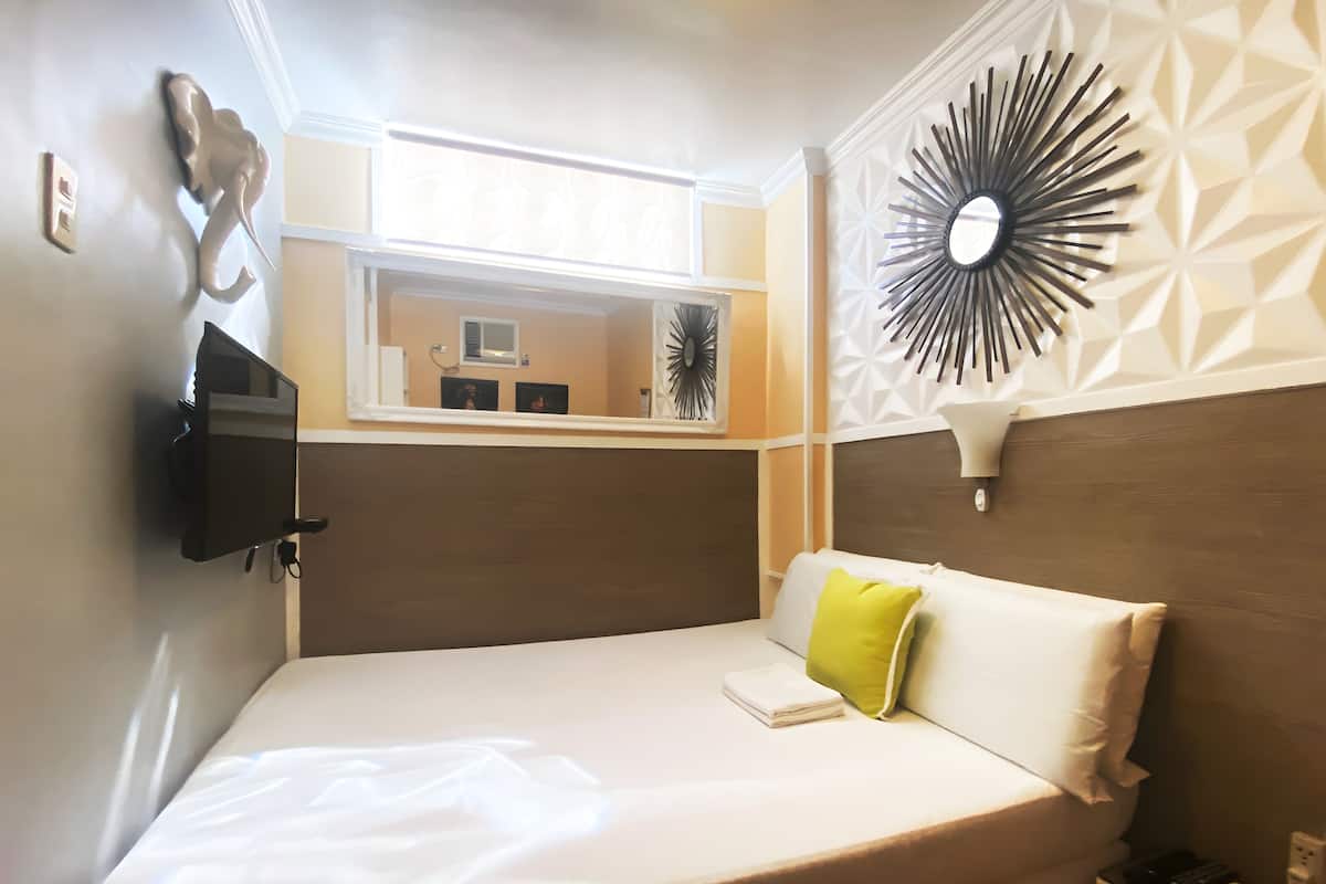 Un lit bien fait, avec une couette blanche et un oreiller vert, un téléviseur fixé au mur, un panneau décoratif mural et une petite table de chevet avec une lampe.