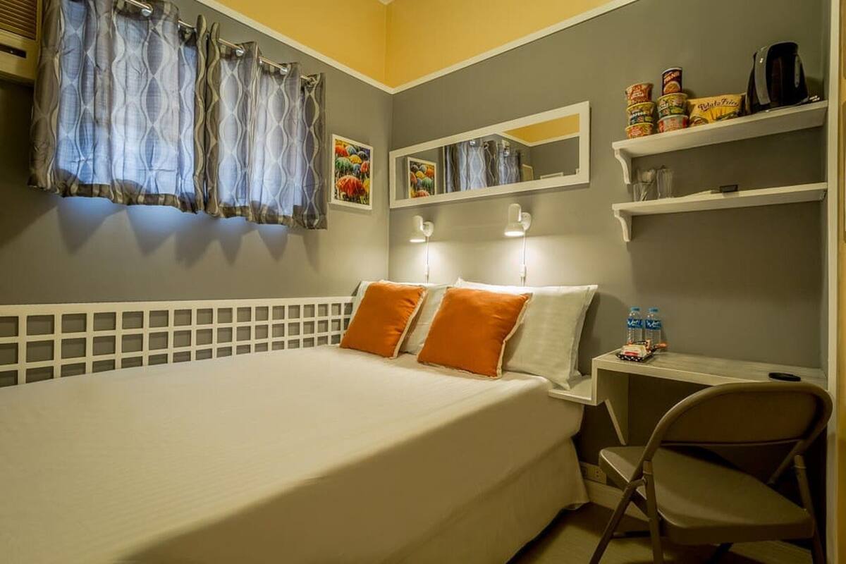 Une chambre à coucher avec un lit, deux oreillers, une chaise, un bureau avec une chaise, une étagère avec des collations, un miroir et des rideaux.