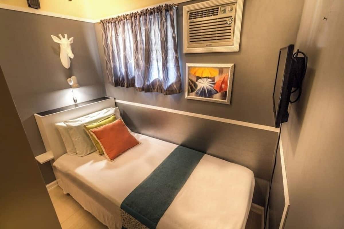 Une chambre d’hôtel de taille réduite, comprenant un lit, une sculpture représentant la tête d’une girafe fixée au mur, un climatiseur mural et un tableau encadré.
