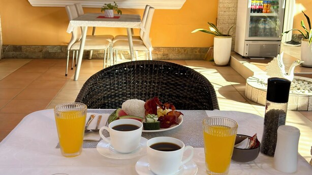Ein Frühstückstisch mit Toast, Eiern, Speck und Obst, dazu Kaffeetassen und Orangensaft.