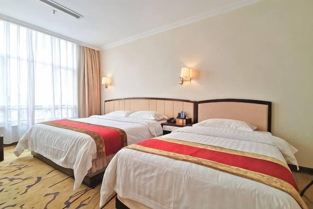 A hotel room with two beds, a large window with curtains, and wall-mounted lamps.