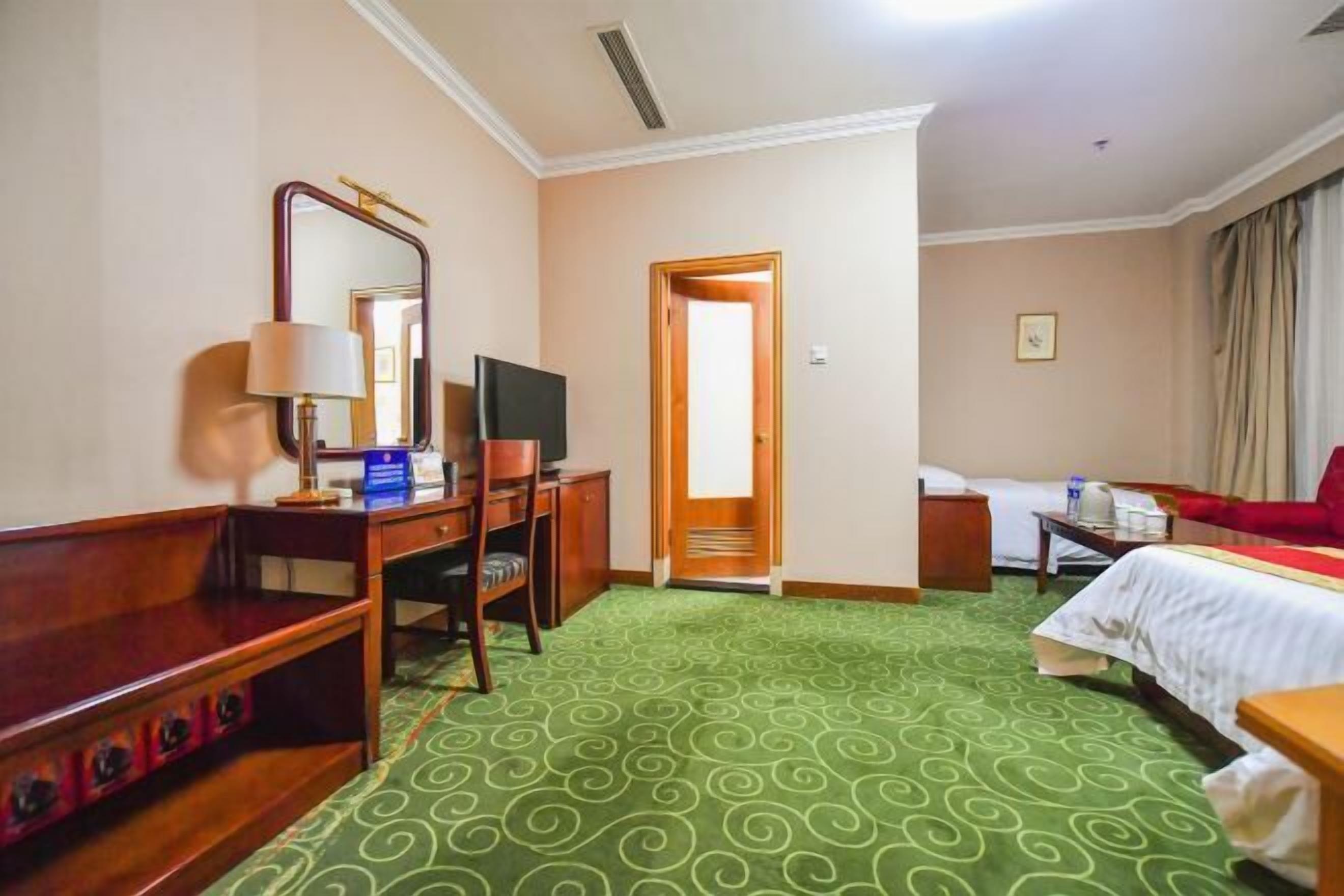 A hotel room with a green patterned carpet, a wooden desk with a lamp, a television, a bed, and a red chair.