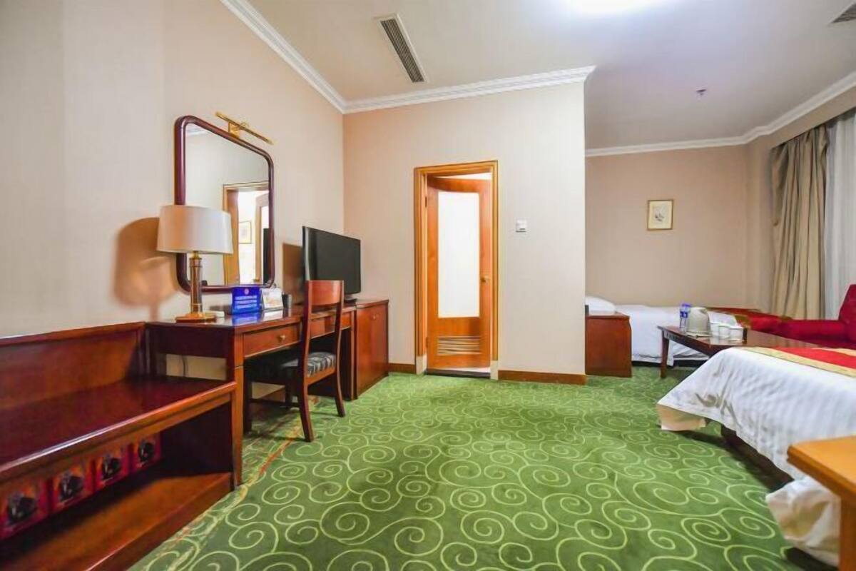 A hotel room with a green patterned carpet, a wooden desk with a lamp, a television, a bed, and a red chair.