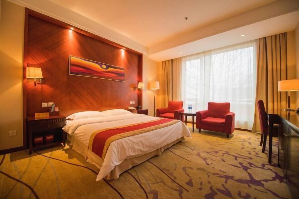 A hotel room with a large bed, two armchairs, a desk, a painting on the wall, and a large window with curtains.