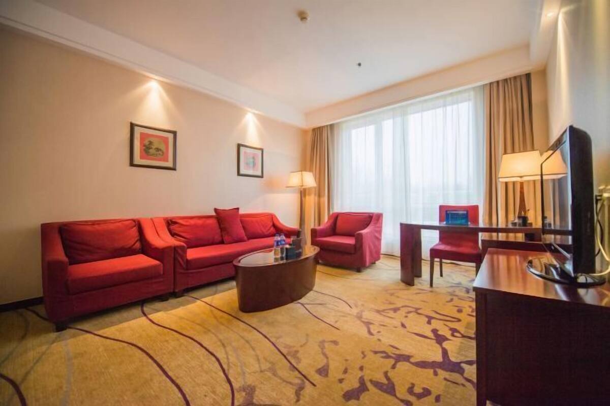 A hotel room with a red sofa set, a coffee table, a TV, and a large window with curtains.