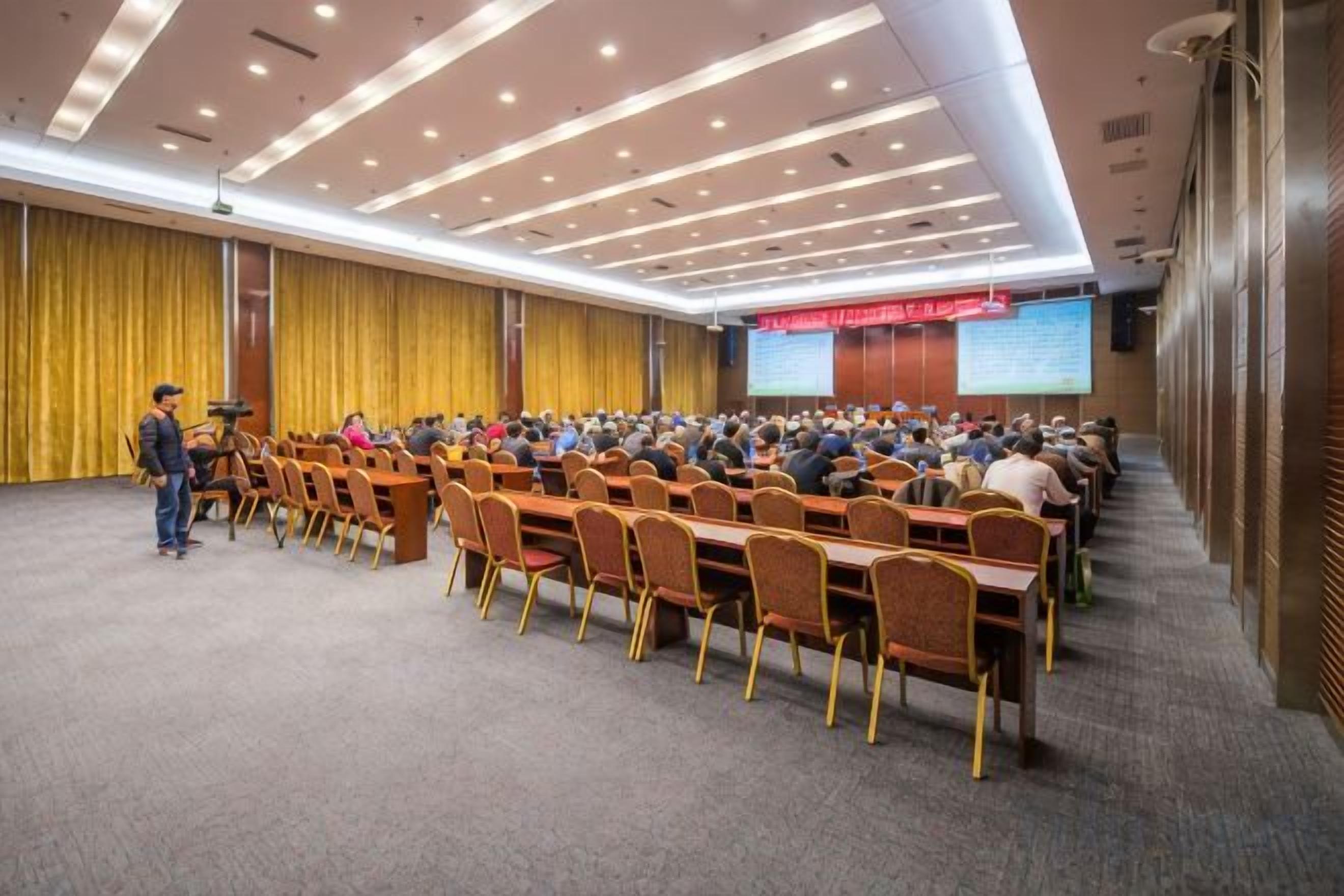 A conference room with rows of chairs, a podium, and a large screen at the front.