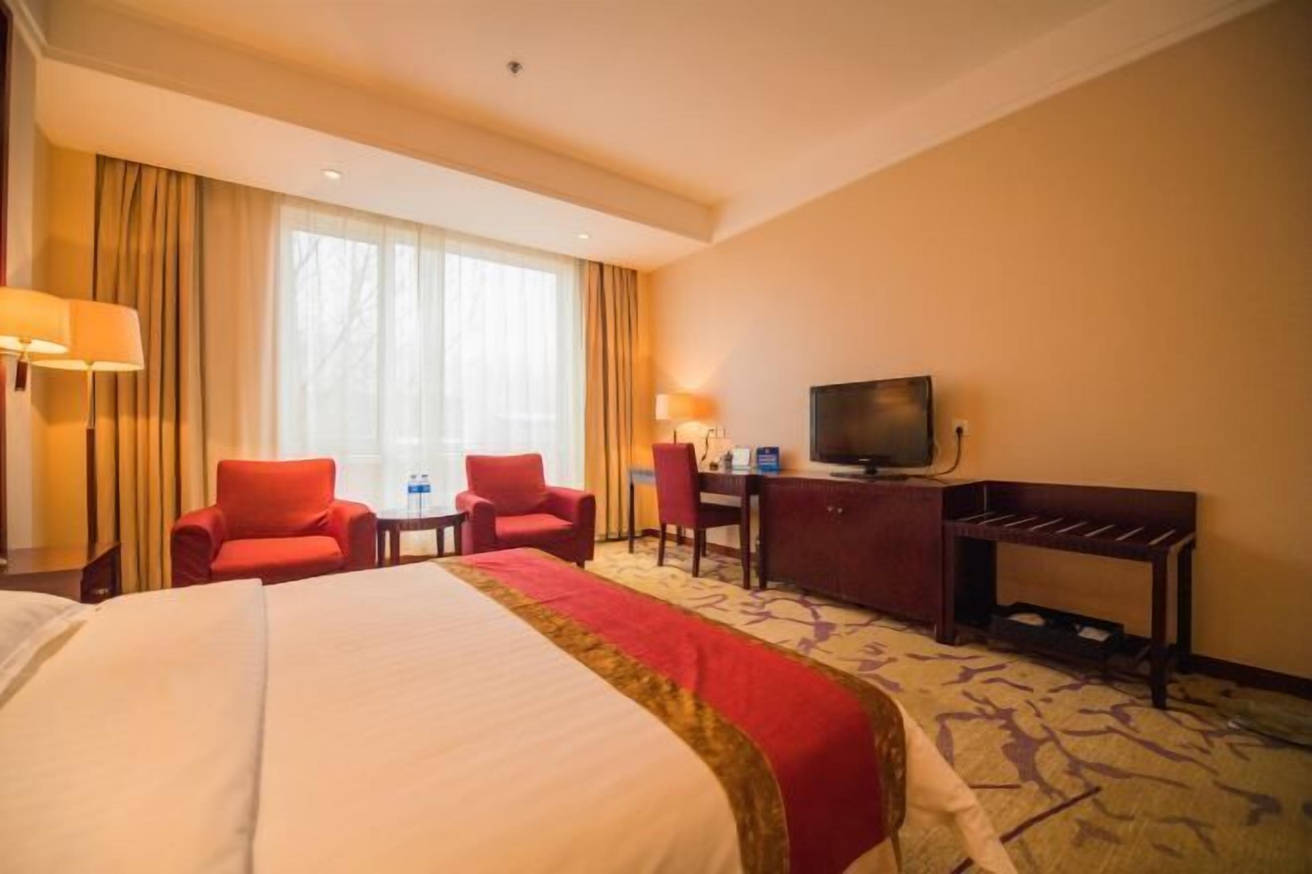 A hotel room with a large bed, two red chairs, a desk with a television, and a window with curtains.