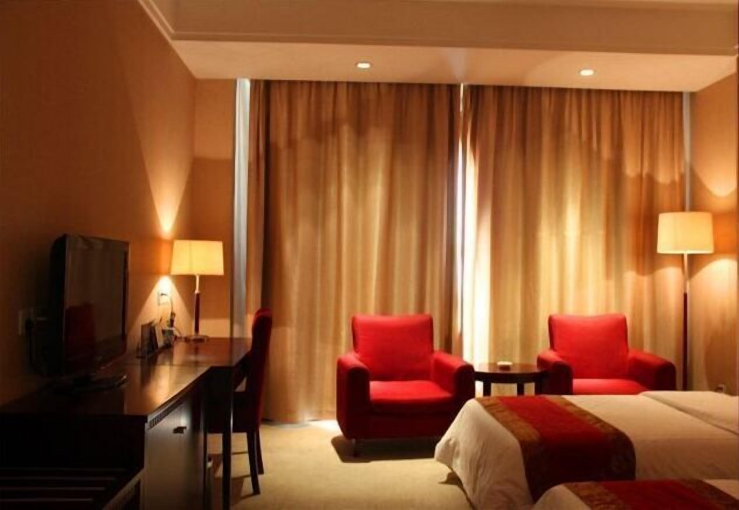 A hotel room with a bed, two red armchairs, a desk with a lamp, and a television.