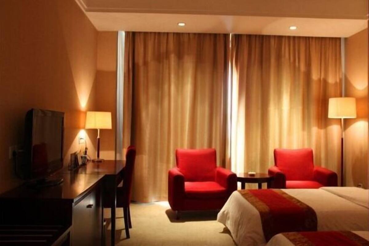 A hotel room with a bed, two red armchairs, a desk with a lamp, and a television.