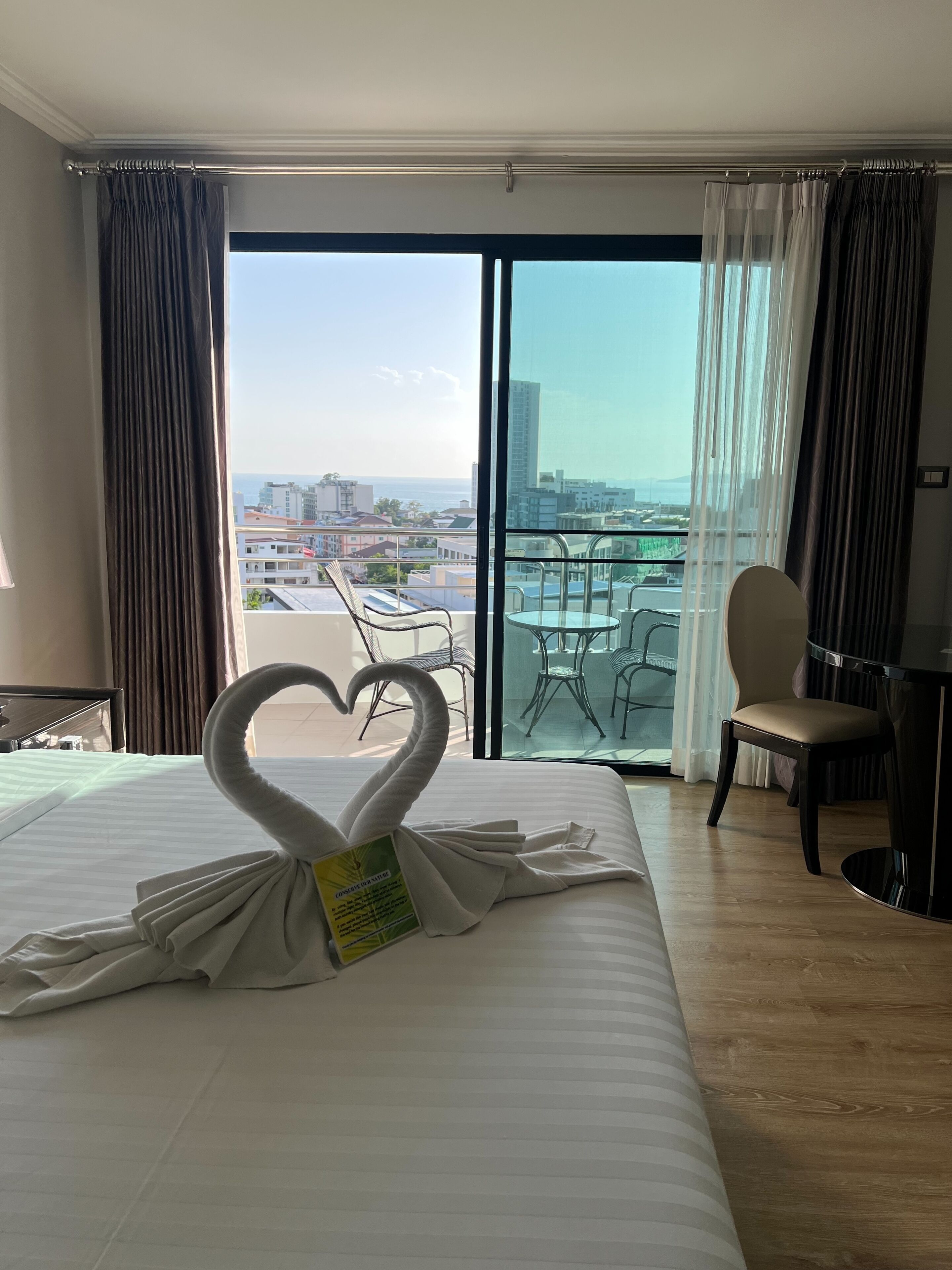 A hotel room with a bed, a heart-shaped towel sculpture, a balcony with a table and chairs, and a view of the cityscape.