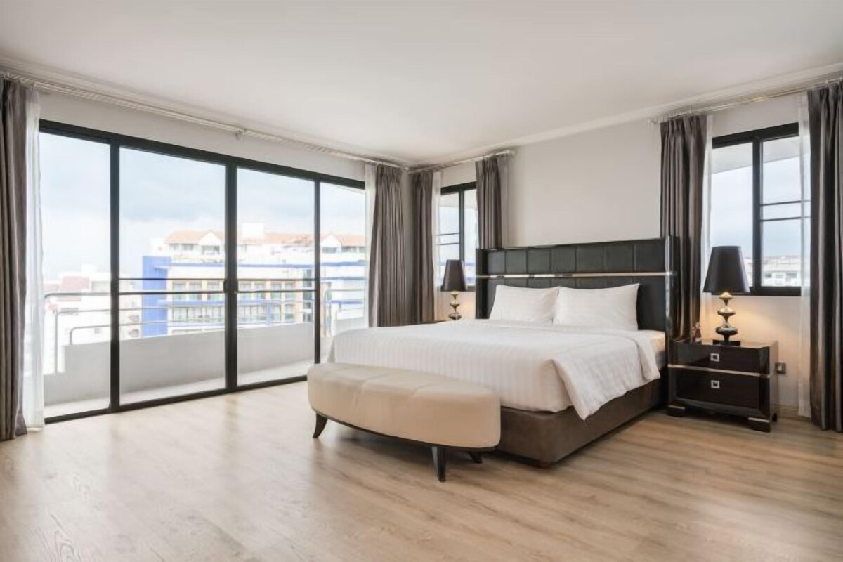 A hotel room with a large bed, a bedside table, a nightstand, and a view of the city through large windows.