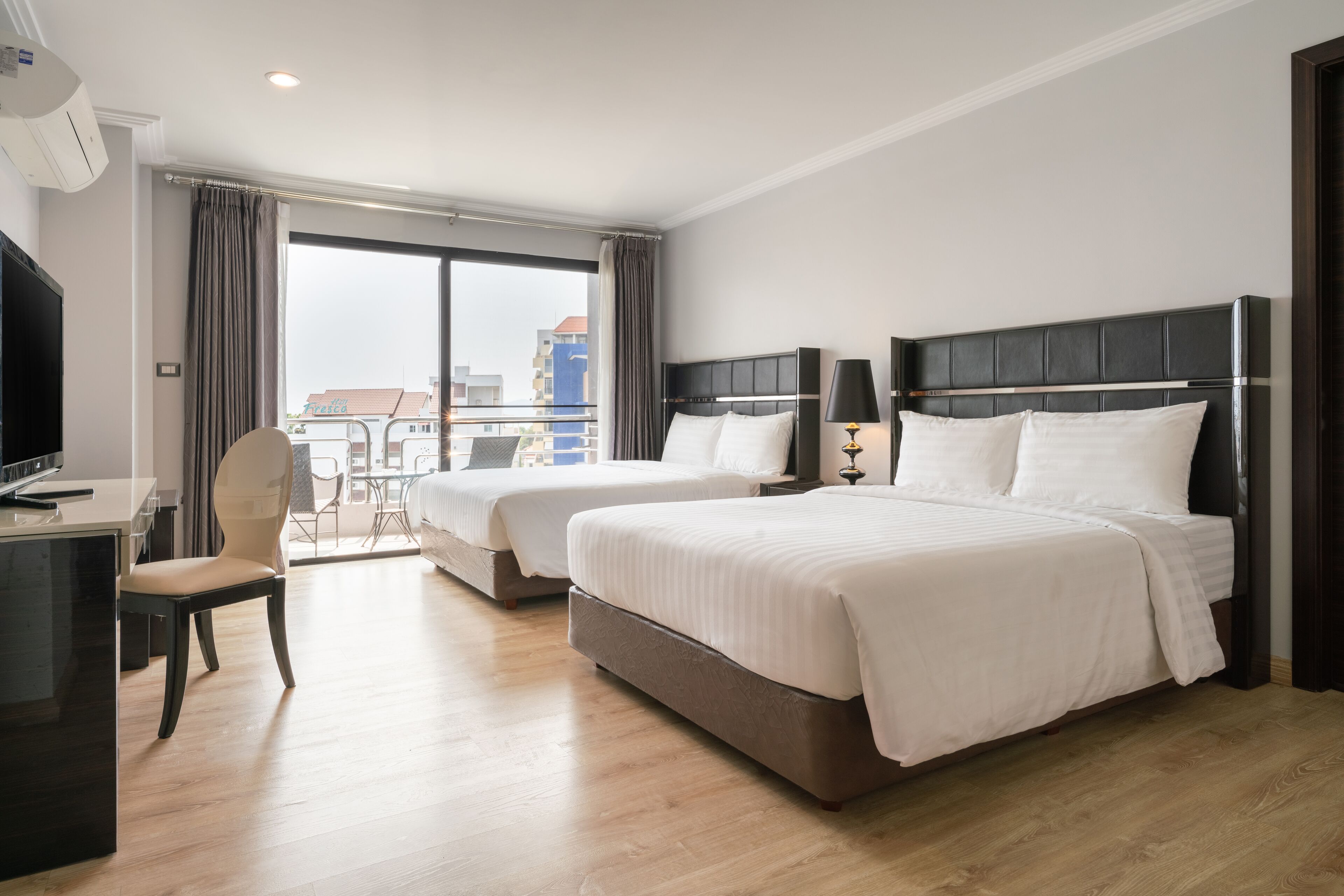 A hotel room with two beds, a desk, and a chair. There is a large window with curtains, and a view of buildings outside.