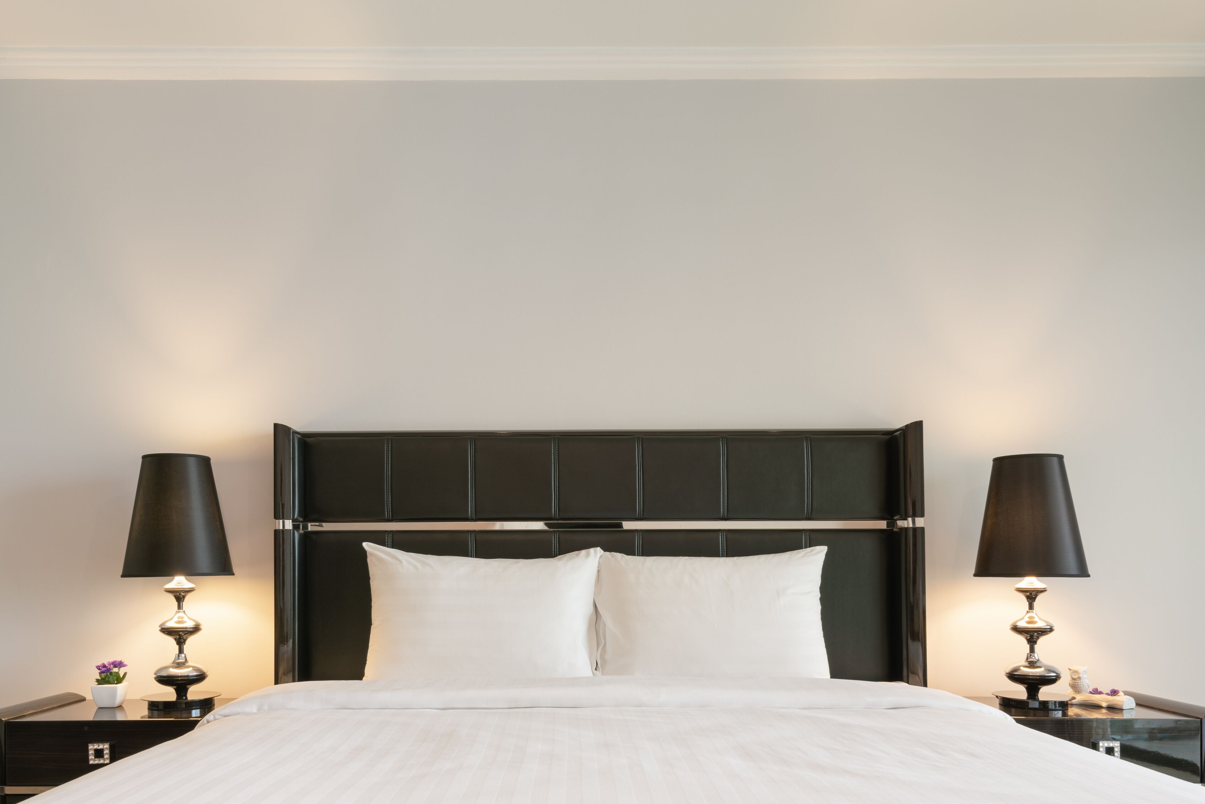 Een netjes opgemaakt bed met witte lakens, met aan beide kanten twee nachtkastjes met lampen, in een kamer met een donker hoofdeinde en effen muren.