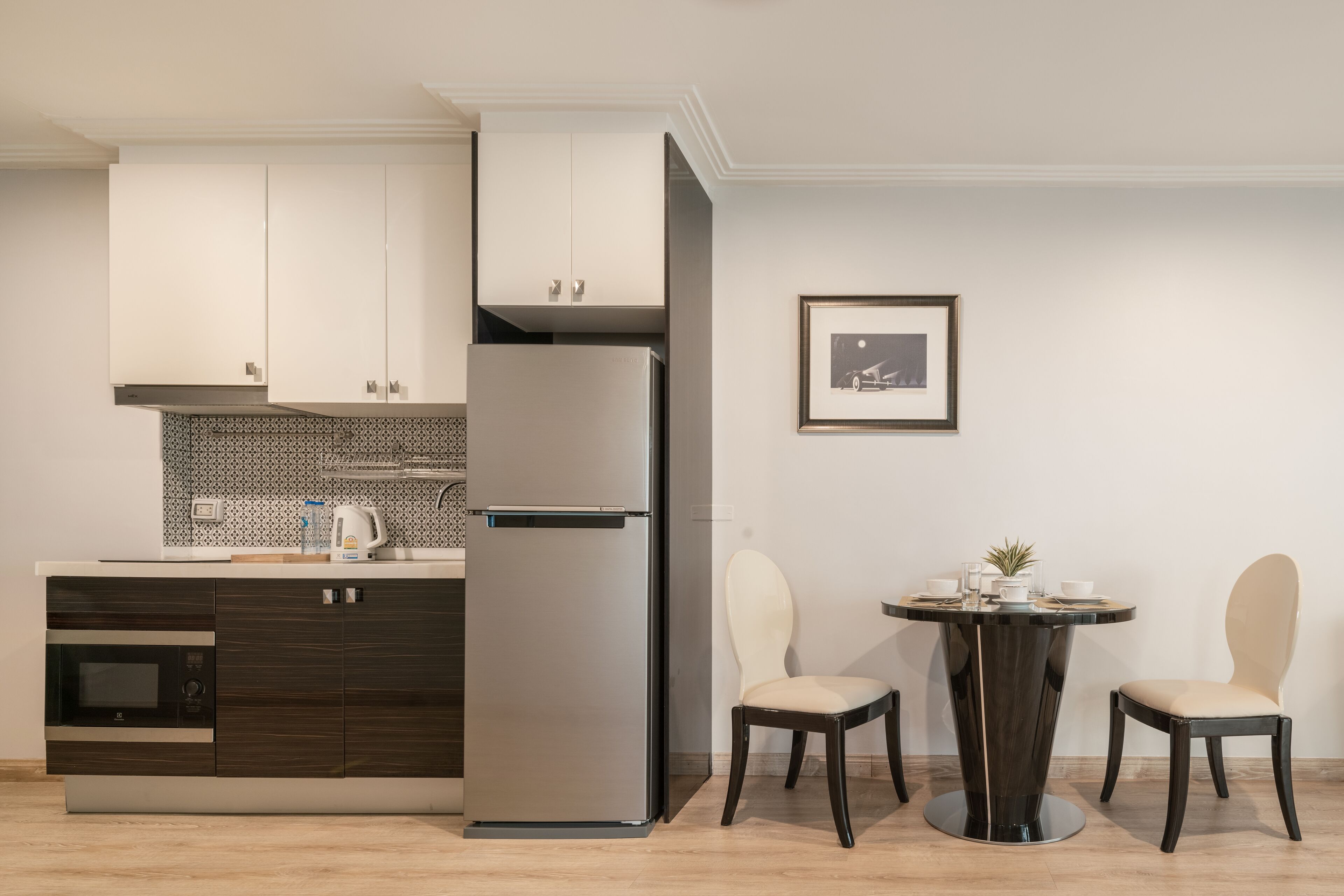 A modern kitchen with a built-in microwave, a refrigerator, and dark wooden cabinets. There is a dining table with two chairs and a framed picture on the wall.